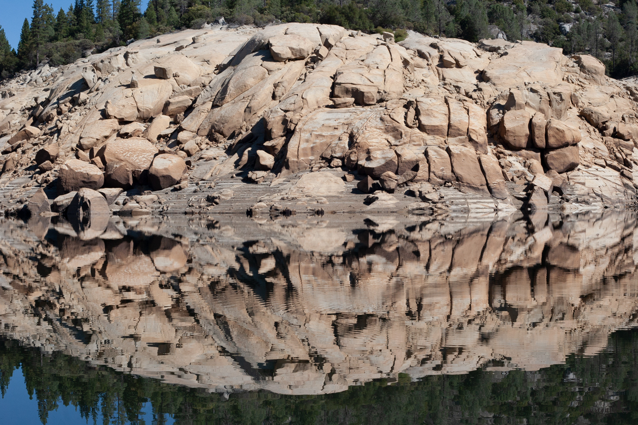 Blue sky, tan boulders and green pine trees reflected in Mammoth Pools Reservoir.