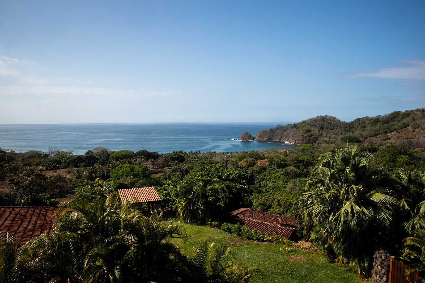 Panoramic view of the Pacific Ocean from Punta Islita, Guanacaste, Costa Rica, featuring lush tropical greenery, terracotta rooftops, and a secluded cove framed by forested hills under a clear blue sky. Days full of sun, serenity, and celebration.
